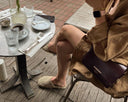 Woman sitting at an outdoor cafe table with a coffee and a flower arrangement, wearing a Burberry handbag.